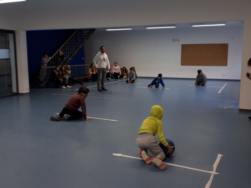 Seis alunos dispostos num campo improvisado de jogo, na sala polivalente da escola, fazendo a sua experimentação de Goalball