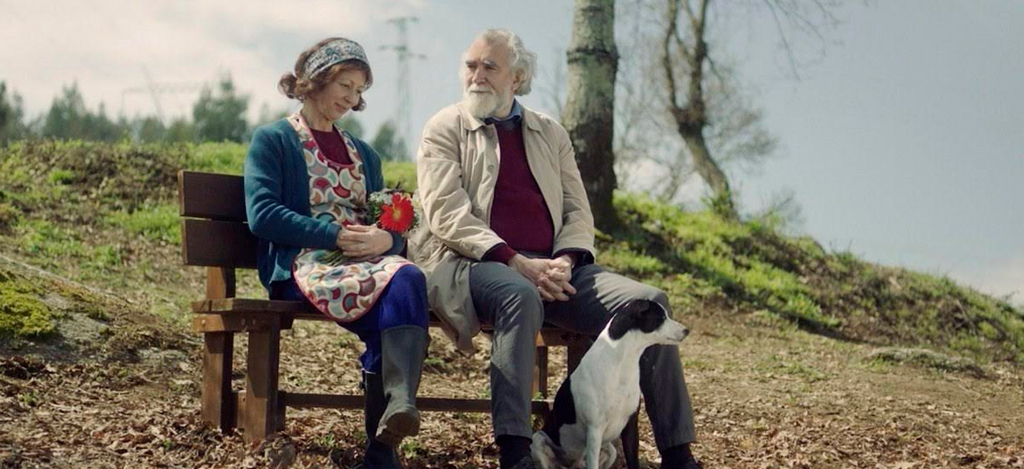 Imagem de uma cena do filme Surdina, mostrando um homem e uma mulher sentados num banco de jardim acompanhados por um cão