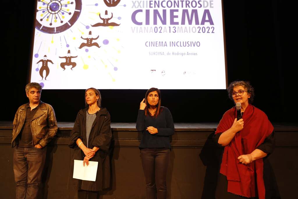 Abertura da sesso de cinema inclusivo. Junto ao palco, e com a tela por trs destacando os XXII Encontros de Cinema de Viana, a investigadora Joslia Neves fala ao pblico. Ao seu lado, a intrprete de Lngua Gestual Portuguesa, um elemento da Organiza