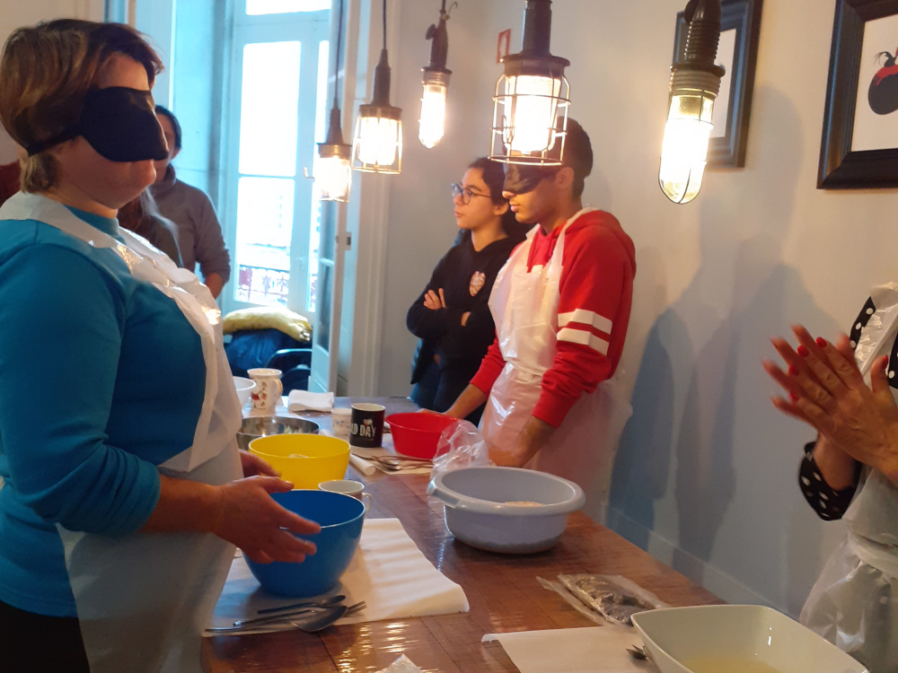Fotografia dos participantes, com os olhos vendados a realizar tarefas de culinária